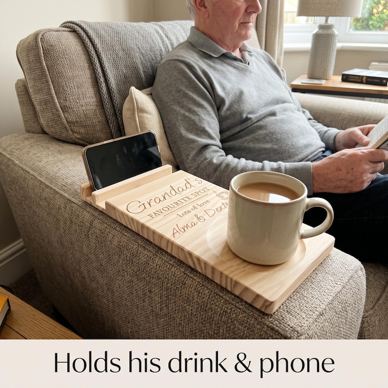 May include: A light-colored wooden armrest tray featuring a slot for a smartphone and a circular indentation for a mug. The surface is engraved with the text "Grandad's FAVOURITE SPOT lots of love Alma & Dexter" and includes a small graphic icon.