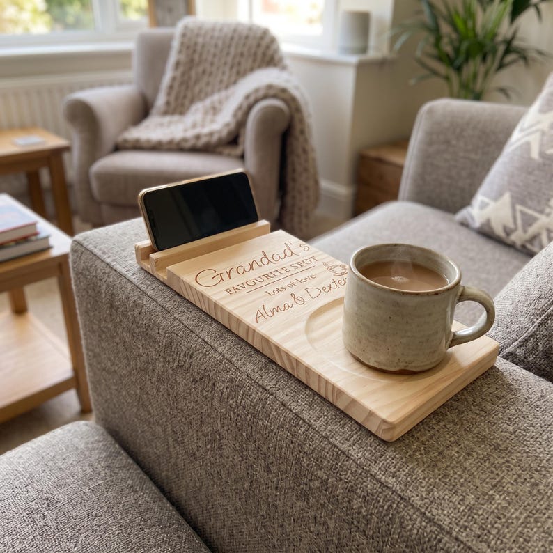 May include: A light-colored wooden sofa arm tray featuring a slot for a smartphone and a circular indentation for a mug. The wood is engraved with the text "Grandad's FAVOURITE SPOT lots of love Alma & Dexter" and a small heart icon.
