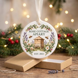 May include: A round white ceramic ornament hanging from a cream ribbon, featuring a printed illustration of a wooden beehive, two bees, and a floral wreath. The text reads ROSE COTTAGE APIARY and EST. 2021. The ornament is presented with a brown box.