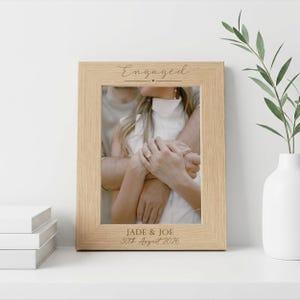May include: A light oak picture frame with the word "Engaged" at the top and the names "Jade & Joe" with the date "30th August 2026" at the bottom. The frame holds a photo of a couple embracing. A white vase with greenery is to the right.