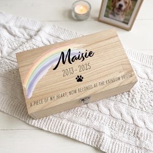 May include: A rectangular light-coloured wooden keepsake box with a metal latch. The lid features a painted pastel rainbow, the name Maisie, the years 2013-2025, a black paw print icon, and the text A PIECE OF MY HEART, NOW BELONGS AT THE RAINBOW BRIDGE.