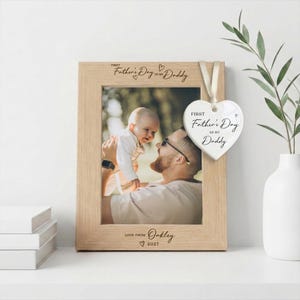 May include: A light wood picture frame with the engraved text "FIRST Father's Day AS MY Daddy" at the top and "LOVE FROM Oakley 2027" at the bottom. A white heart-shaped ceramic ornament hangs from the frame with the text "FIRST Father's Day AS MY Daddy".