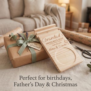 May include: A rectangular light-colored wooden stand with a circular indentation and a grooved slot. The wood is engraved with the text: Grandad's FAVOURITE SPOT, Lots of love, Alma & Dexter x, and a small coffee cup icon.