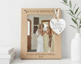 May include: A light wood picture frame with the engraved text TODAY MY BRIDESMAID FOREVER MY BEST FRIEND and 11.7.26. A white heart-shaped ceramic ornament hangs from the frame with the same text and small black heart icons printed on it.