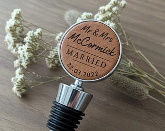 May include: A bottle stopper featuring a circular tan leather insert with black engraved text reading Mr & Mrs McCormick MARRIED 22.03.2022. The stopper has a polished silver metal base and a black ribbed rubber seal for securing bottles.