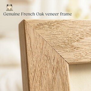 May include: A light brown wooden frame corner featuring a mitered joint and a visible natural wood grain texture. The text "Genuine French Oak veneer frame" is printed above the frame, accompanied by a small illustration of a frog.