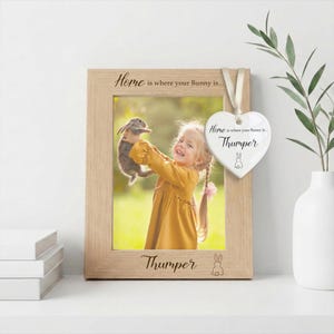 May include: A light wood picture frame with the text Home is where your Bunny is... and Thumper engraved on the frame. A white heart-shaped ceramic ornament hangs from the frame with the text Home is where your Bunny is... Thumper.