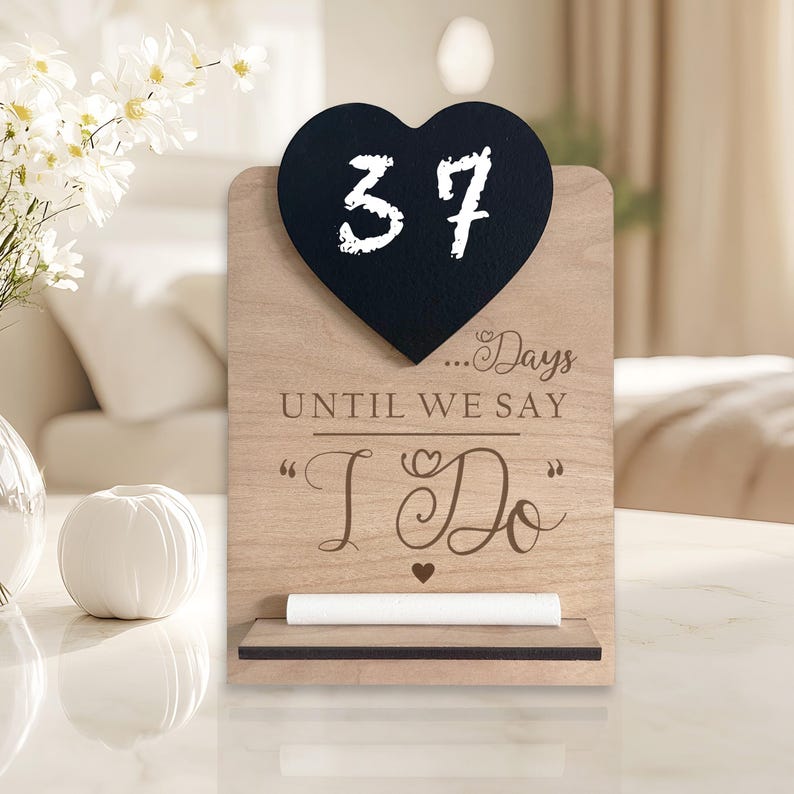 以下が含まれることがあります： 「37...Days Until We Say "I Do"」と書かれた黒いハート型のチョークボードが付いた木製のカウントダウンサイン。チョークボードは、チョークを置くための小さな棚が付いた木製スタンドに取り付けられています。