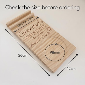 May include: A rectangular wooden tray with a grooved slot and a circular indentation. The tray features engraved text reading "Grandad's FAVOURITE SPOT", "Lots of love", and "Alma & Dexter x", plus a coffee cup icon. Dimensions are 26cm by 12cm.
