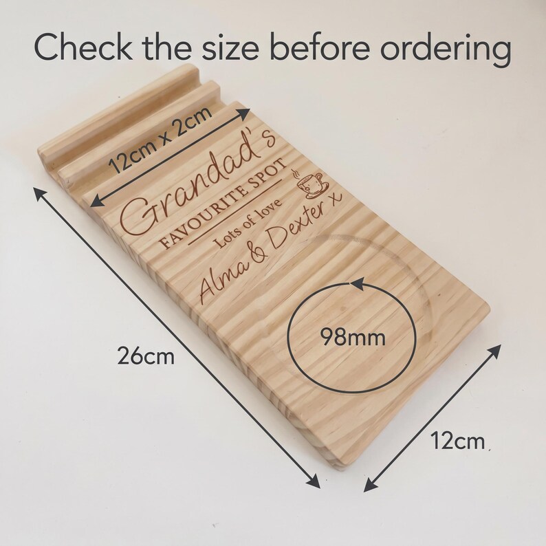 May include: A rectangular wooden tray with a grooved slot and a circular indentation. The tray features engraved text reading "Grandad's FAVOURITE SPOT", "Lots of love", and "Alma & Dexter x", plus a coffee cup icon. Dimensions are 26cm by 12cm.