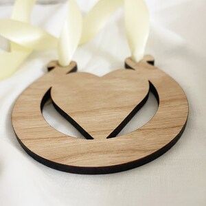 May include: A wooden ornament shaped like a horseshoe with a heart cut out in the center. The ornament is light brown and has a dark brown border. It is attached to a pale yellow ribbon.