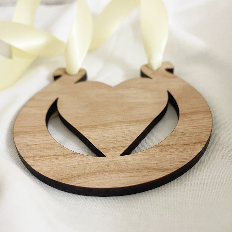 May include: A wooden ornament shaped like a horseshoe with a heart cut out in the center. The ornament is light brown and has a dark brown border. It is attached to a pale yellow ribbon.