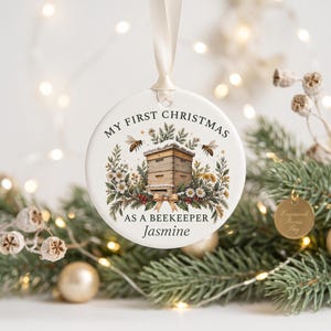 May include: A round white ceramic ornament hanging from a cream ribbon. The center features a graphic of a wooden beehive surrounded by flowers, greenery, and two bees. The text reads: My First Christmas As A Beekeeper, with the name Jasmine below.