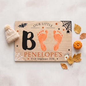 以下が含まれることがあります： 「OUR LITTLE」の文字、大きな黒い「B」、2つのオレンジ色の赤ちゃんの足跡、「PENELOPE'S First Halloween 2026」の文字が描かれた長方形の木製看板。デザインには黒いコウモリ、クモの巣、星、小さな幽霊のイラストが含まれています。