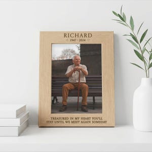 May include: A light oak picture frame with the name "RICHARD" and dates "1947-2024" engraved at the top. The frame contains a photograph of an elderly man seated on a bench. The bottom of the frame reads "TREASURED IN MY HEART YOU'LL STAY UNTIL WE MEET AGAIN SOMEDAY."