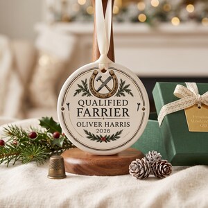 May include: A round white ceramic ornament hanging from a cream ribbon. The design features a horseshoe, a hammer, and a nail, with the text "QUALIFIED FARRIER," "OLIVER HARRIS," and "2026" printed in black, surrounded by green pine sprigs.