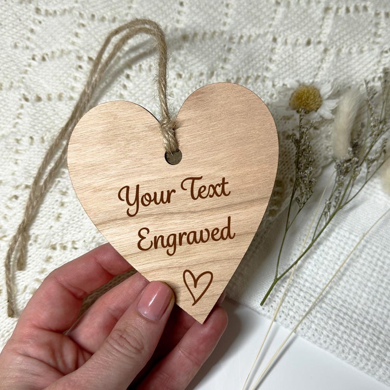 May include: A wooden heart-shaped ornament with the text "Your Text Engraved" and a small heart symbol. The ornament is light brown and has a hole at the top for hanging with a twine string. Decorative dried flowers are in the background.