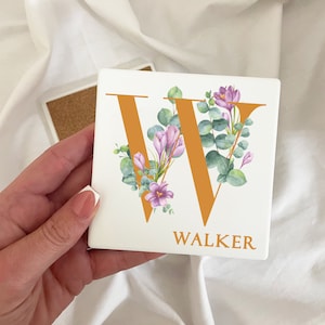 May include: A white ceramic coaster with a floral design featuring a large orange letter W surrounded by green leaves and purple flowers. The text "WALKER" is printed below the letter.