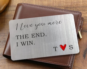May include: A rectangular brushed silver metal wallet insert card featuring the black engraved text I Love you more THE END. I WIN. with the letters T and S separated by a small red heart symbol. The card is placed on a brown leather wallet.