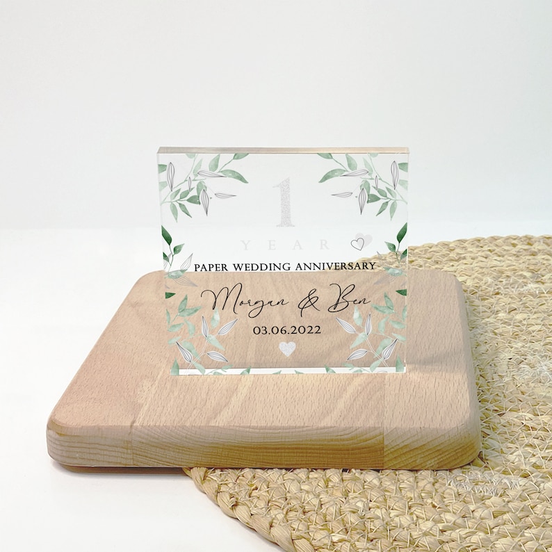May include: A clear acrylic block with the text "1 YEAR PAPER WEDDING ANNIVERSARY" and the names "Morgan & Ben" with the date 03.06.2022. The block is decorated with green leaves and rests on a wooden base.