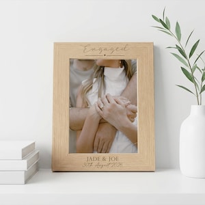 May include: A light oak picture frame with the word "Engaged" at the top and the names "Jade & Joe" with the date "30th August 2026" at the bottom. The frame contains a photo of a couple embracing. A white vase with greenery is to the right.