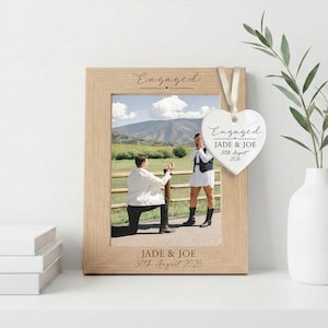 May include: A light wood picture frame featuring the engraved text Engaged and JADE & JOE 30th August 2026. A white heart-shaped ceramic ornament hangs from the frame with the text Engaged, JADE & JOE, and 30th August 2026 printed in black ink.
