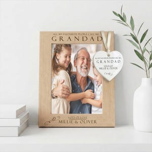 May include: A light wood picture frame with engraved text reading ALL MY FAVOURITE PEOPLE CALL ME GRANDAD and LOTS OF LOVE MILLIE & OLIVER. A white heart-shaped ceramic ornament hangs from the frame with matching printed text and a cream ribbon.