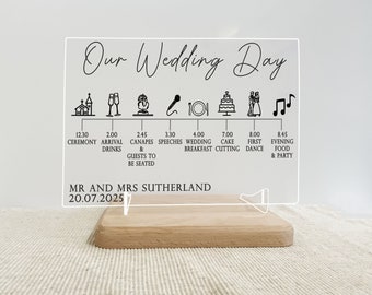 May include: A clear acrylic sign with a black outline and text that reads "Our Wedding Day". The sign lists the wedding day timeline with icons and times for the ceremony, arrival, canapes, speeches, wedding breakfast, cake cutting, first dance, and evening food and party. The sign also includes the names "MR AND MRS SUTHERLAND" and the date "20.07.2025".