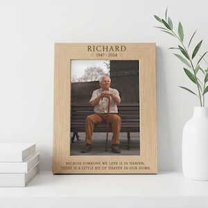 May include: A wooden picture frame with the name "RICHARD" and dates "1947-2024" engraved at the top. The frame holds a photograph of an older man sitting on a bench. The bottom of the frame has the text: "BECAUSE SOMEONE WE LOVE IS IN HEAVEN, THERE IS A LITTLE BIT OF HEAVEN IN OUR HOME."