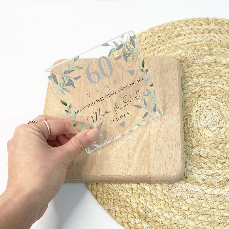 May include: A clear acrylic plaque with silver text and floral accents. The plaque commemorates a 60th wedding anniversary, with the words "60 YEARS DIAMOND WEDDING ANNIVERSARY" and the names "Mum & Del" and the date "25.11.1964".