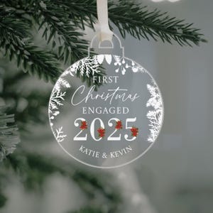 May include: Clear acrylic Christmas ornament with the text "FIRST Christmas ENGAGED 2025" and the names "KATIE & KEVIN". The ornament is round with a white floral design and red berry accents. A white ribbon is attached for hanging.