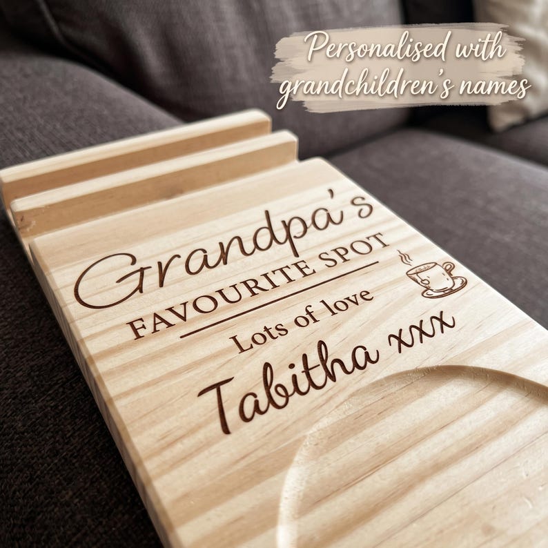 May include: A light-colored wooden coaster and phone stand featuring engraved text that reads Grandpa's FAVOURITE SPOT, Lots of love, and Tabitha xxx, accompanied by a small coffee cup icon and a circular indentation for a beverage container.