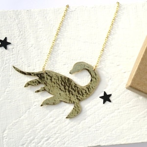 May include: A gold necklace with a hammered metal pendant shaped like a mythical creature. The necklace is packaged in a brown box with a black bow and the text "Metal Dove".