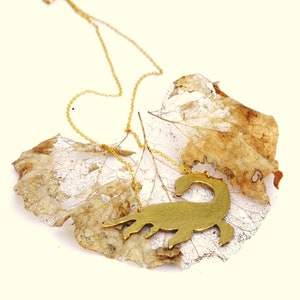 May include: A gold necklace with a scorpion pendant. The pendant is a cutout design and is hanging on a delicate gold chain. The necklace is displayed on a dried leaf.