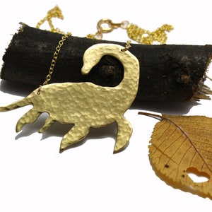 May include: Gold necklace featuring a hammered brass pendant shaped like a stylized dinosaur. The necklace is displayed on a piece of driftwood with a dried leaf.