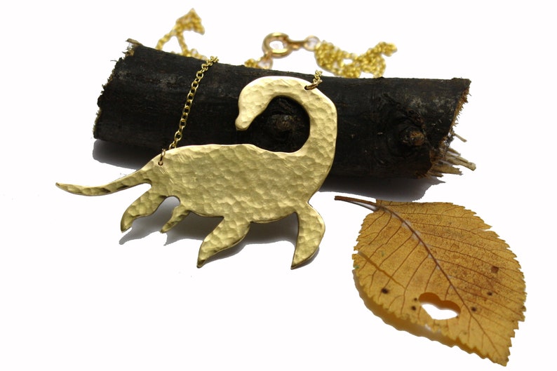 May include: Gold necklace featuring a hammered brass pendant shaped like a stylized dinosaur. The necklace is displayed on a piece of driftwood with a dried leaf.