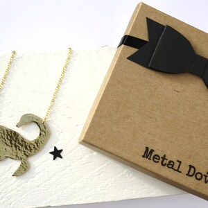 May include: A gold necklace with a hammered metal pendant shaped like a swan. The necklace is packaged in a brown box with a black bow and the words "Metal Dove" printed on the box.