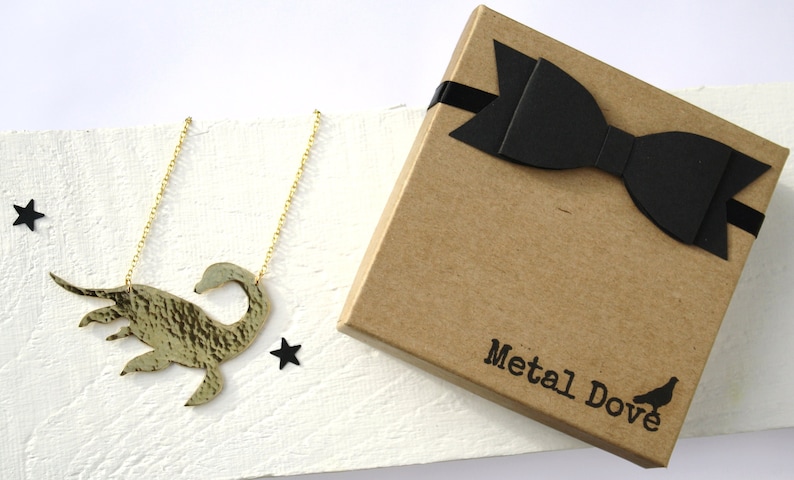 May include: A gold necklace with a hammered metal pendant shaped like a swan. The necklace is packaged in a brown box with a black bow and the words "Metal Dove" printed on the box.