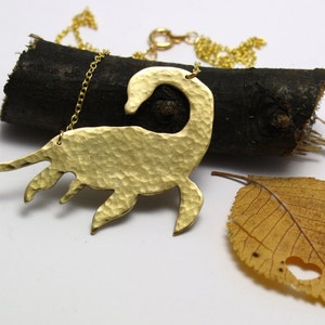 May include: A gold necklace with a hammered brass pendant shaped like a Loch Ness Monster. The pendant is hanging on a gold chain and is resting on a piece of wood.