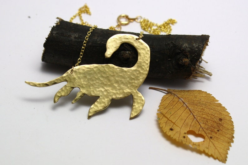 May include: A gold necklace with a hammered brass pendant shaped like a Loch Ness Monster. The pendant is hanging on a gold chain and is resting on a piece of wood.
