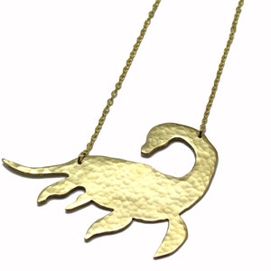 May include: A gold necklace with a hammered, textured pendant shaped like a Loch Ness Monster. The pendant is large and detailed, with a long neck and a small head.