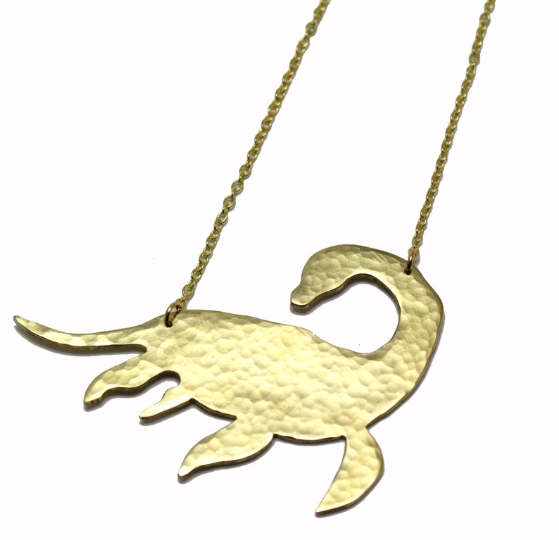 May include: A gold necklace with a hammered, textured pendant shaped like a Loch Ness Monster. The pendant is large and detailed, with a long neck and a small head.
