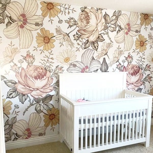 Puede incluir: Una cuna blanca con un fondo de papel tapiz floral. El papel tapiz presenta un patrón floral a gran escala con flores rosas, amarillas y marrones.