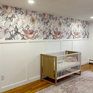May include: A white crib with a wooden frame and a floral patterned wallpaper behind it. A pink and white rug is on the floor in front of the crib.