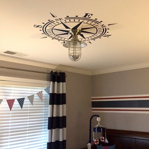 May include: A nautical themed nursery with a compass ceiling decal, a striped curtain, and a crib with a "dream big" pillow.