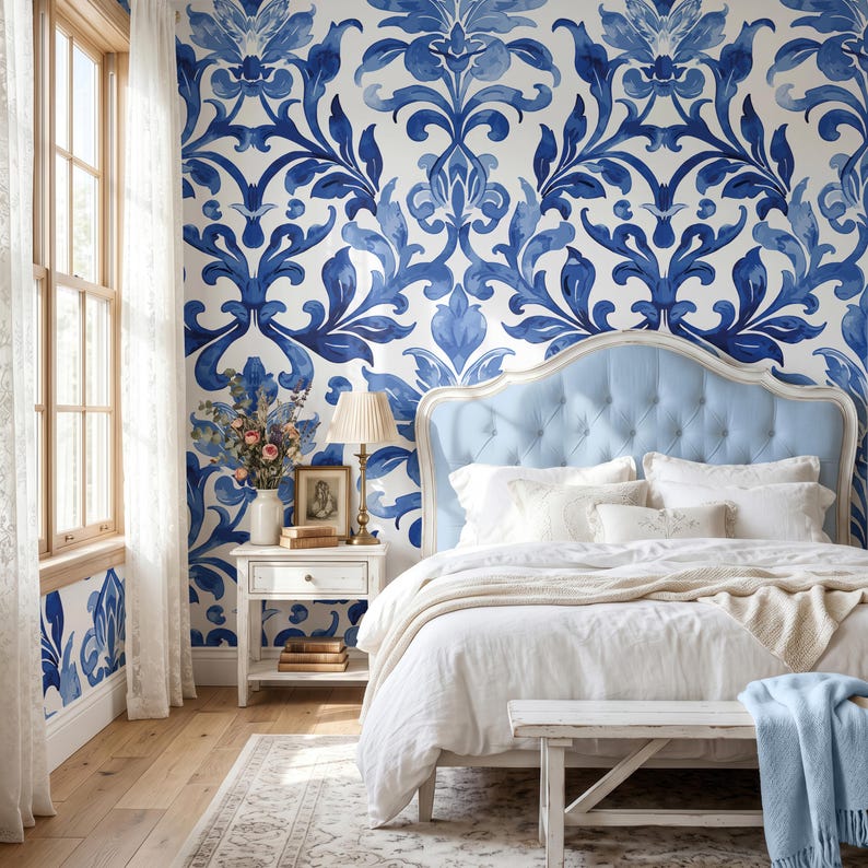 May include: A bedroom featuring a light blue tufted headboard, white bedding, and a white wooden nightstand. The wall is covered in wallpaper with a large-scale blue and white damask pattern. A lamp, a framed picture, and books sit on the nightstand.