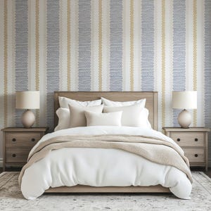 Op de afbeelding: Een slaapkamer met een bed met wit beddengoed, een beige sprei en een houten hoofdbord. Twee bijpassende nachtkastjes met lampen flankeren het bed. Het behang heeft verticale strepen in blauw, goud en wit.
