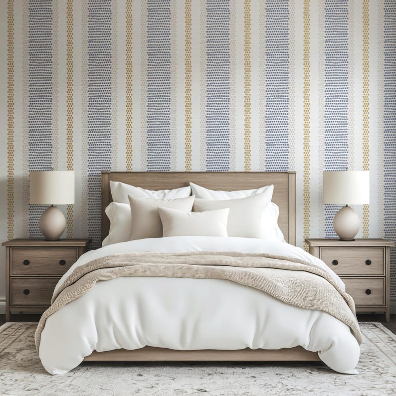 Op de afbeelding: Een slaapkamer met een bed met wit beddengoed, een beige sprei en een houten hoofdbord. Twee bijpassende nachtkastjes met lampen flankeren het bed. Het behang heeft verticale strepen in blauw, goud en wit.