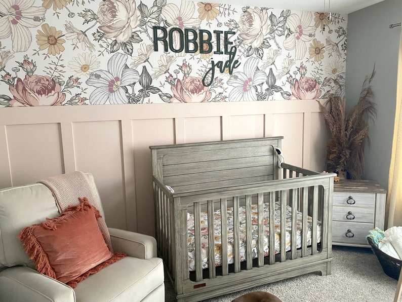 May include: A nursery room with a crib, a rocking chair, and a dresser. The walls are covered in floral wallpaper with the names "Robbie" and "Jade" written on the wall.