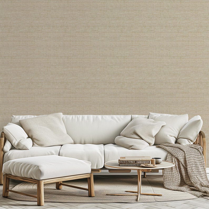 May include: A living room scene featuring a white sofa with several beige pillows and a matching ottoman. A small round table holds books and a cup. A beige throw blanket and rug complete the neutral color scheme.